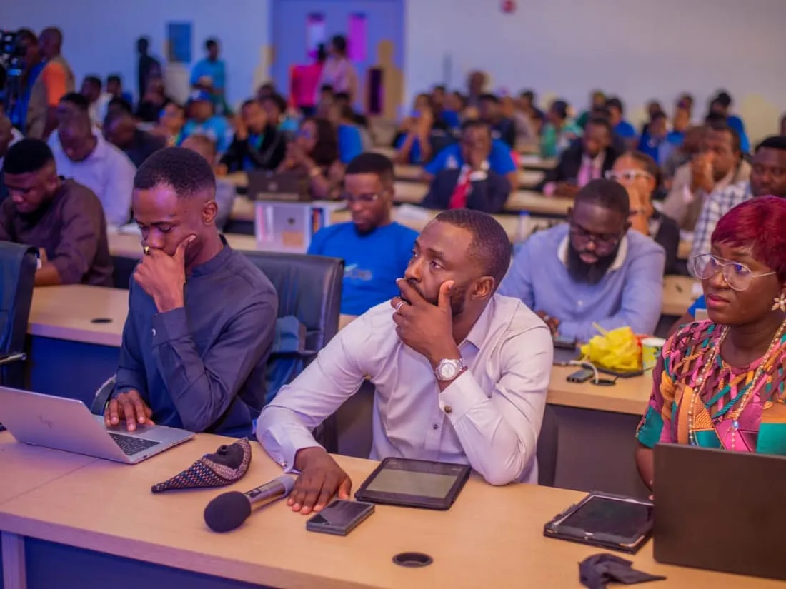 Participants listening at desks with laptops and tablets during a training session.
