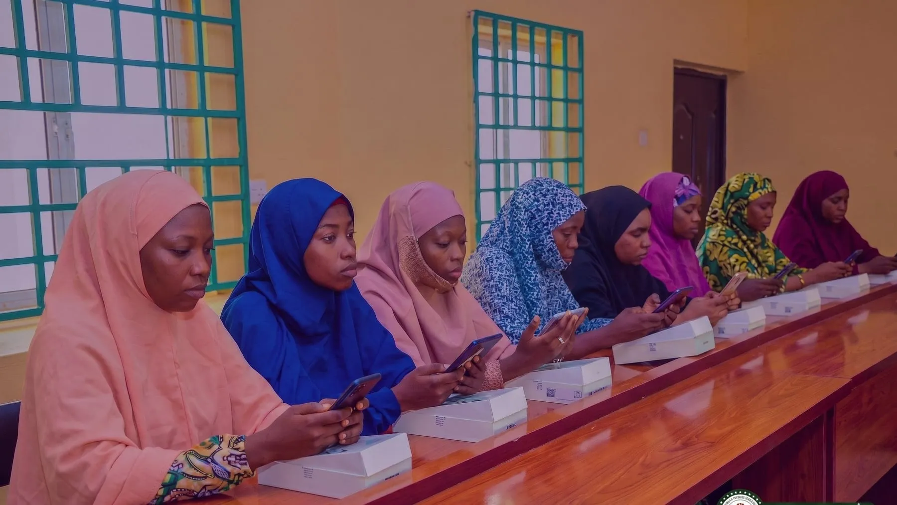 A row of adolescent girls in hijabs seated at a long desk inside the Sokoto Digital Village hub, each using a newly issued smartphone with the device boxes still in front of them.