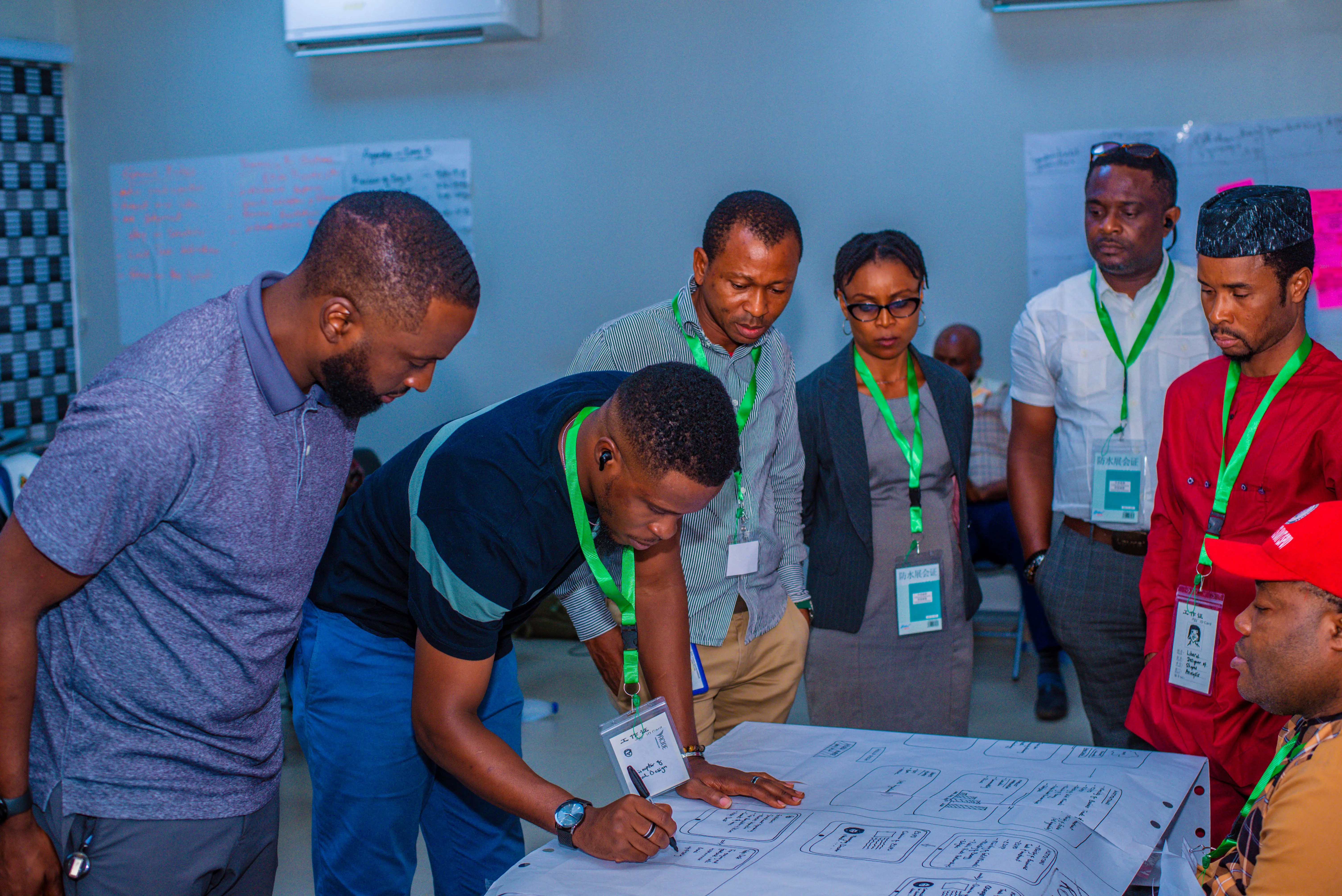 Participants gathered around a table in a Bragi workshop, one sketching a service-flow diagram on a large sheet while colleagues look on, with brainstorming flip-charts on the wall behind.